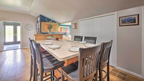 A dining area with a table set for six, wooden chairs, a clock on the wall, and a kitchen in the background. - Loft Unplugged w/ Outdoor Space in Amish Country! (Middlefield)