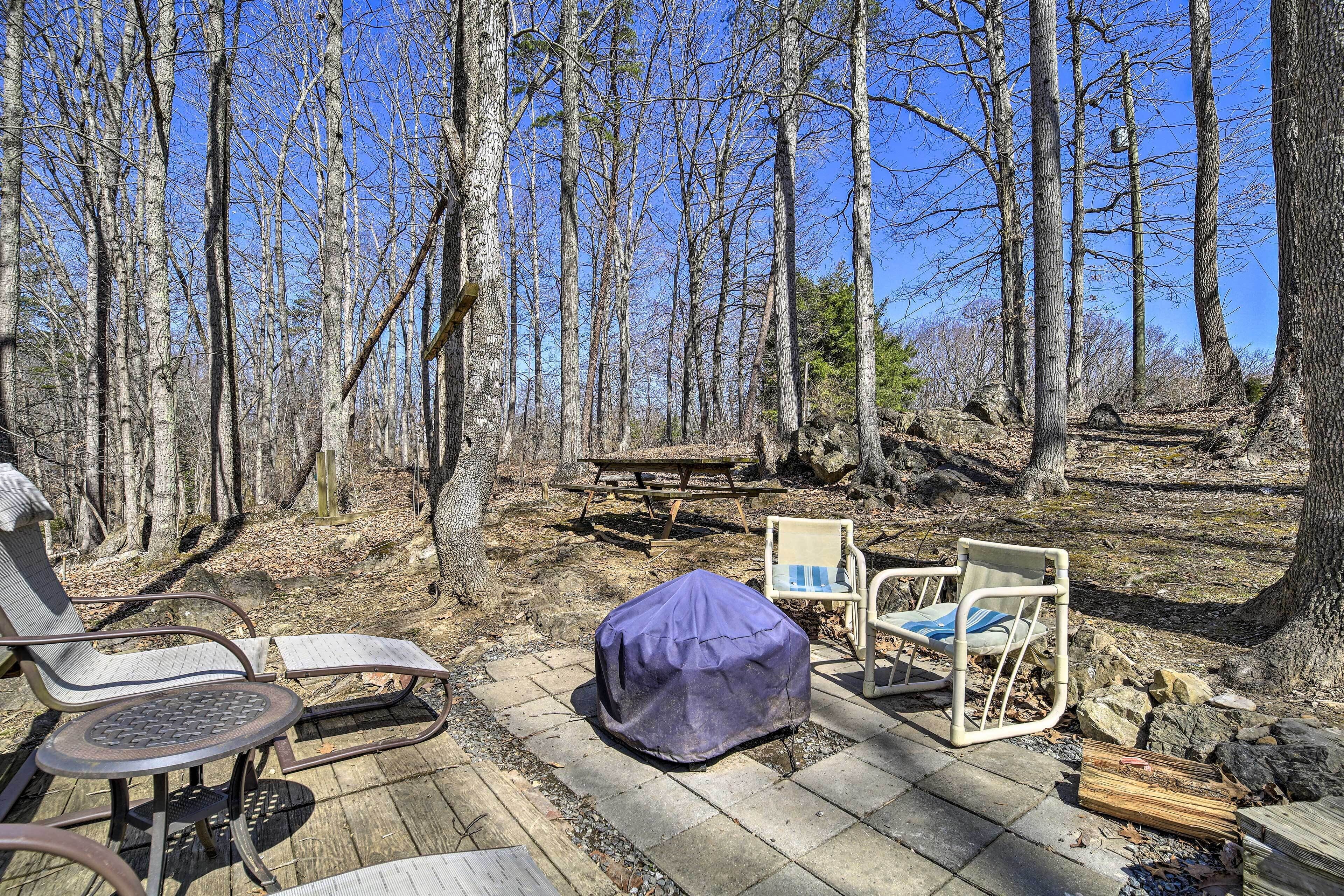A wooden deck with two covered chairs, a picnic table, and a fire pit in a forested area.