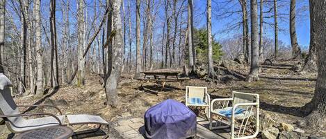 A wooden deck with two covered chairs, a picnic table, and a fire pit in a forested area.