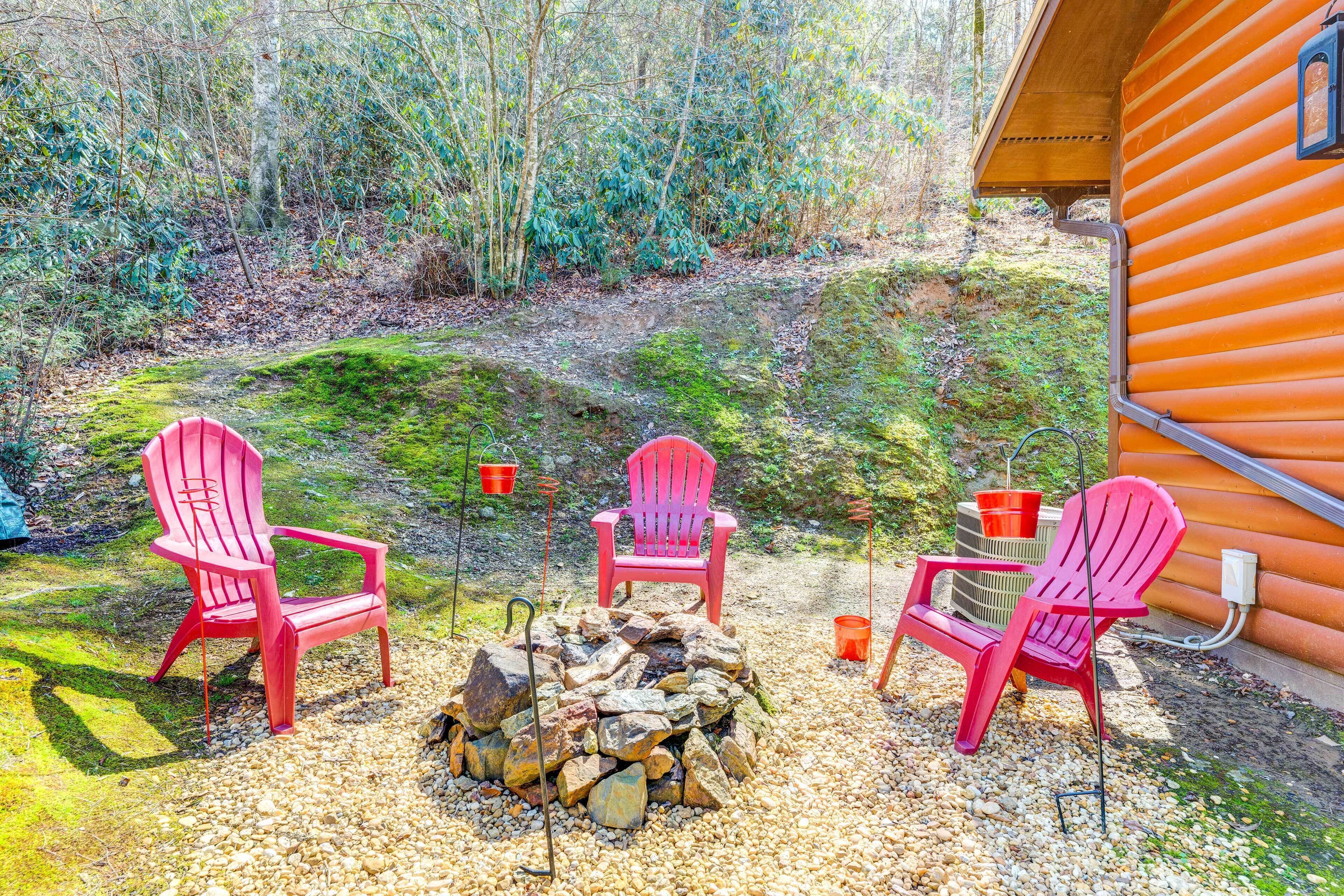 A cabin with a fire pit and three Adirondack chairs in a forested area.
