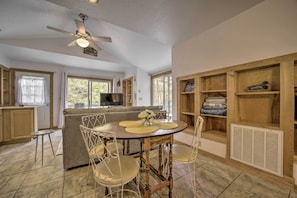 A dining area with a round table, chairs, and a ceiling fan. There is a kitchen area with cabinets and a window.