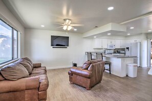 Una sala moderna con un sofá y un sillón de cuero marrón, una cocina con gabinetes y electrodomésticos blancos, y un ventilador de techo.