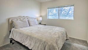 Un dormitorio con cama, mesita de noche, lámpara y ventana con vistas a árboles.