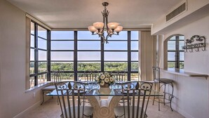 A dining area with a glass table, metal chairs, and a chandelier. Large windows offer a view of greenery. - Sunny Getaway: Community Amenities, Walk to Beach! (Marco Island)