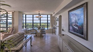 A spacious living room with a large window, a dining area, and a painting on the wall. - Sunny Getaway: Community Amenities, Walk to Beach! (Marco Island)