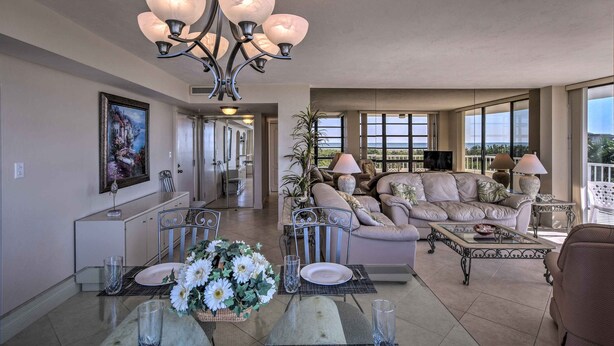 A spacious living room with a dining area, a chandelier, a sofa set, and large windows offering a view. - Sunny Getaway: Community Amenities, Walk to Beach! (Marco Island)