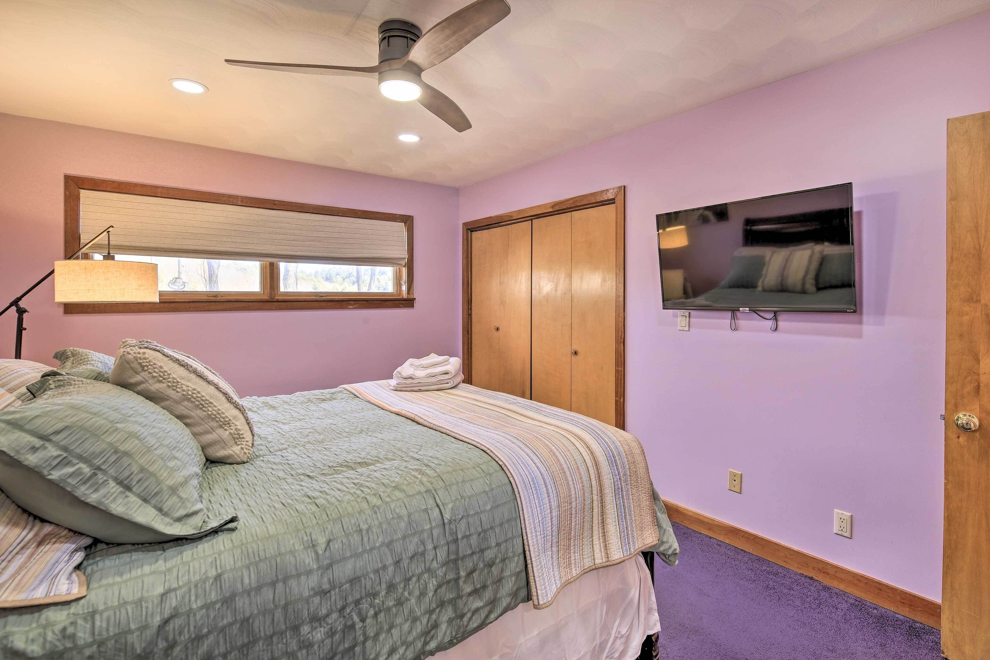 A bedroom with a bed, a ceiling fan, a window with blinds, a television on the wall, and a wooden wardrobe.