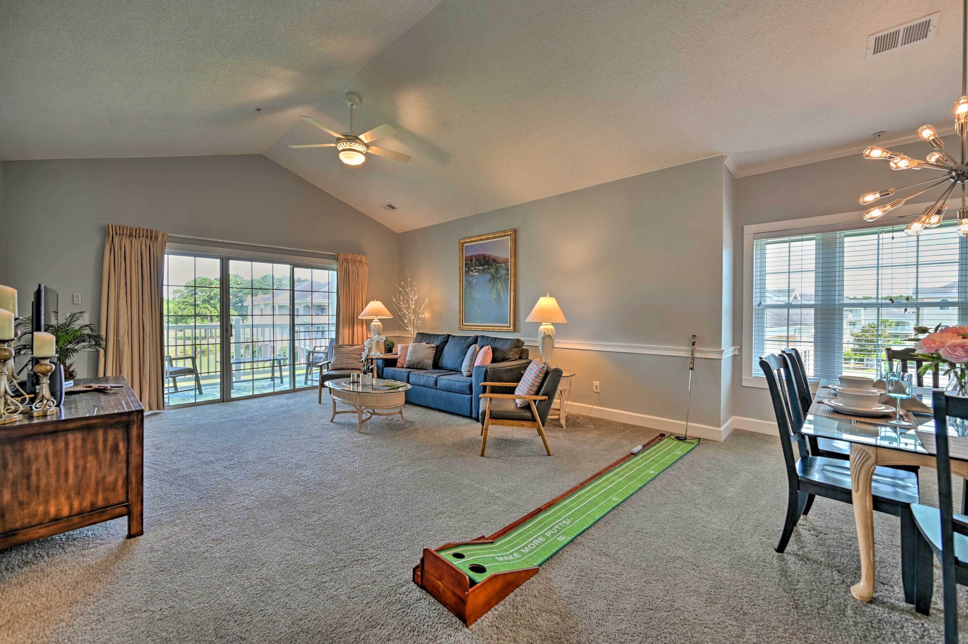 A spacious living room with a large window, a ceiling fan, a dining area with a table set for two, and a shuffleboard table.