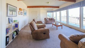 A spacious living room with a high ceiling, built-in bookshelves, two sofas, and a large window offering a view of the outdoors. - Fishermen's Paradise in Coos Bay: Walk to Beach! (Coos Bay)