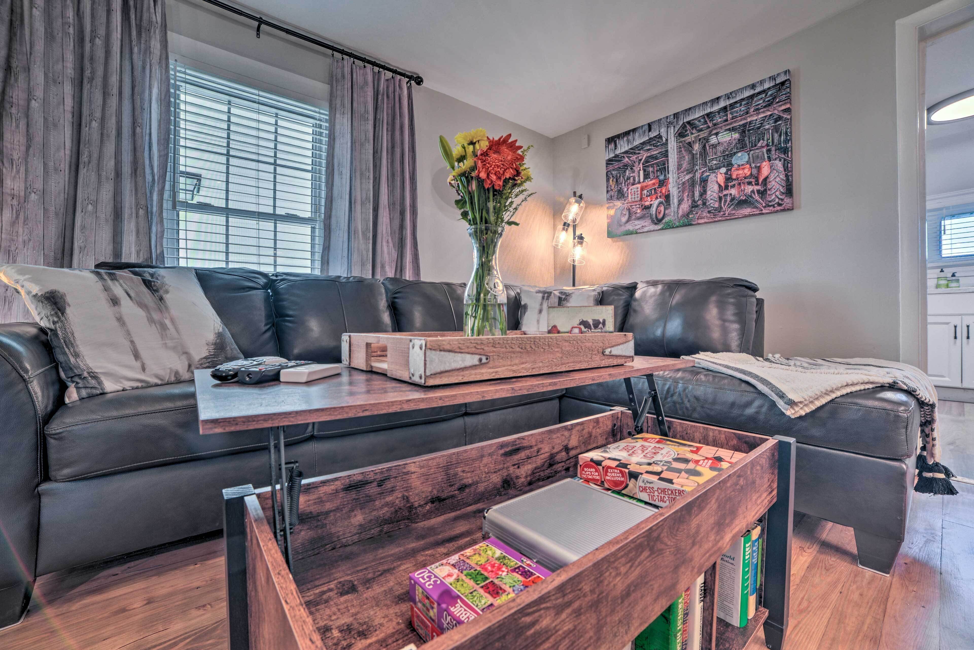 Un salon moderne avec un canapé en cuir foncé, une table basse en bois ornée de livres et d’un vase de fleurs, et un tableau accroché au mur.