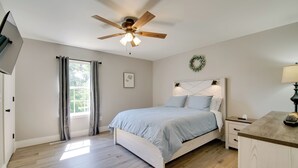 Une chambre à coucher comprenant un lit, un ventilateur de plafond, une fenêtre avec des rideaux, une table de chevet et une commode.