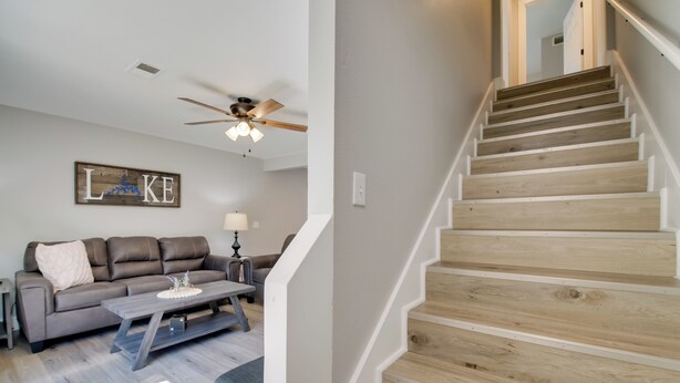 Un escalier avec des marches en bois et des rampes blanches, un canapé gris agrémenté d’un coussin décoratif, une table basse en bois et un ventilateur de plafond avec des lampes.