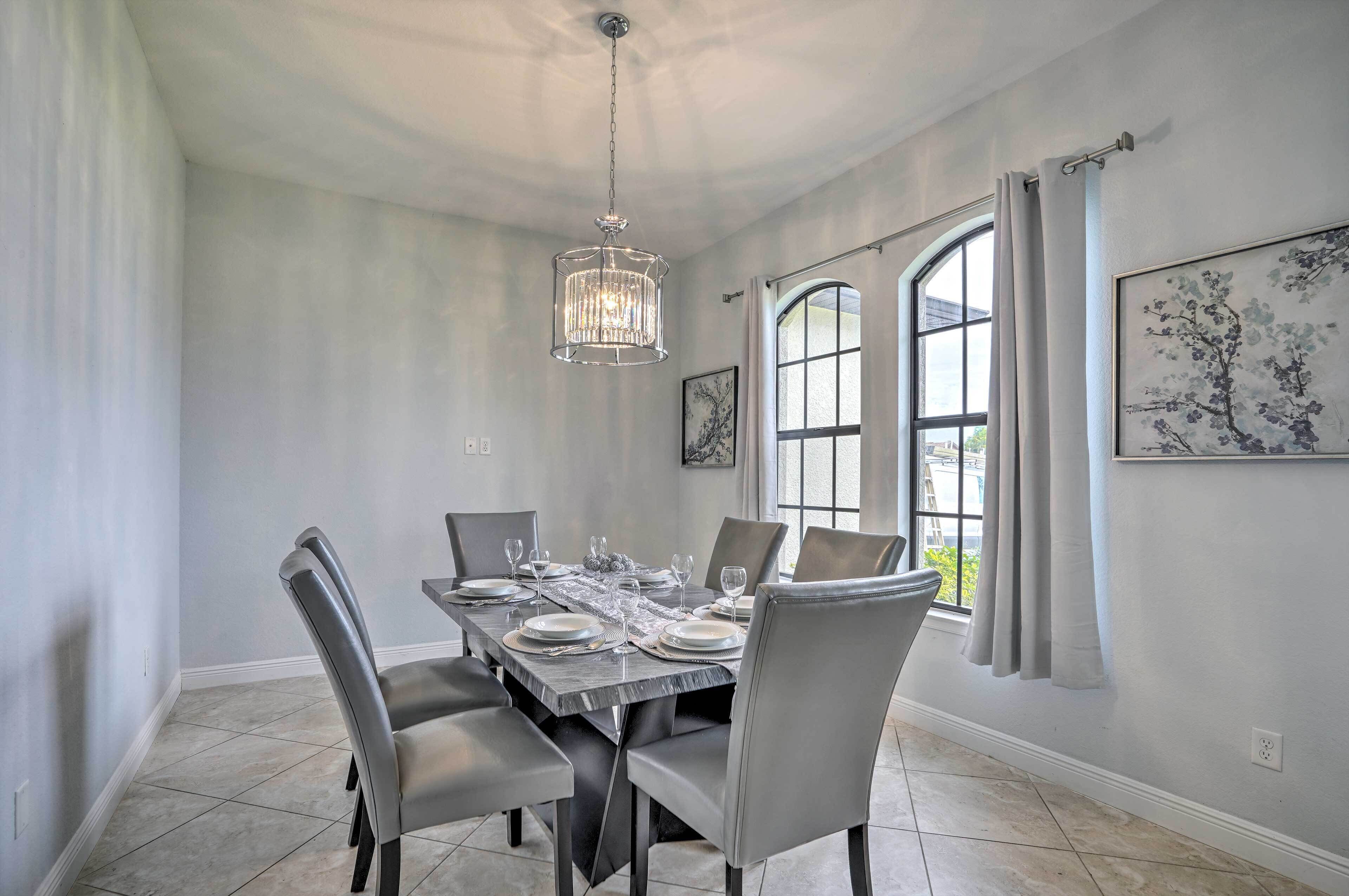 A dining room with a large table set for six, a chandelier, and a window with curtains.