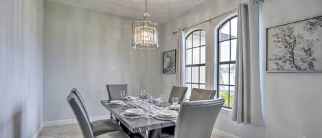 A dining room with a large table set for six, a chandelier, and a window with curtains.