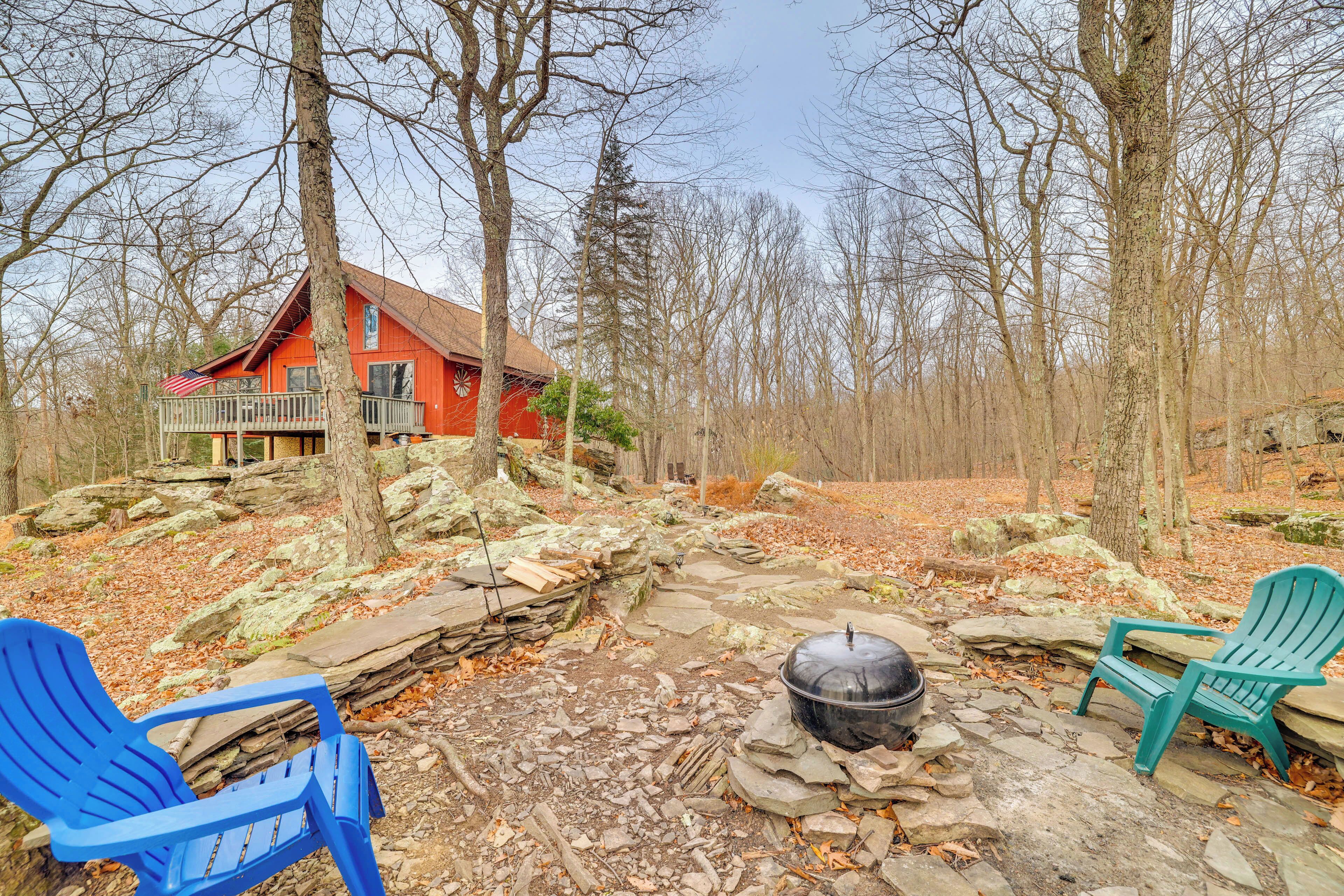 A rustic cabin with a stone fire pit and Adirondack chairs in a wooded area.