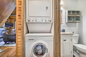 A compact laundry room with a front-loading washing machine, a wooden door, and a small shelf with toiletries. - Secluded Cresco Cabin w/ Deck + Forest Views! (Cresco)