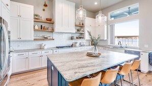 Una cocina moderna con armarios blancos, una isla grande con taburetes de bar, iluminación colgante y vistas al exterior.