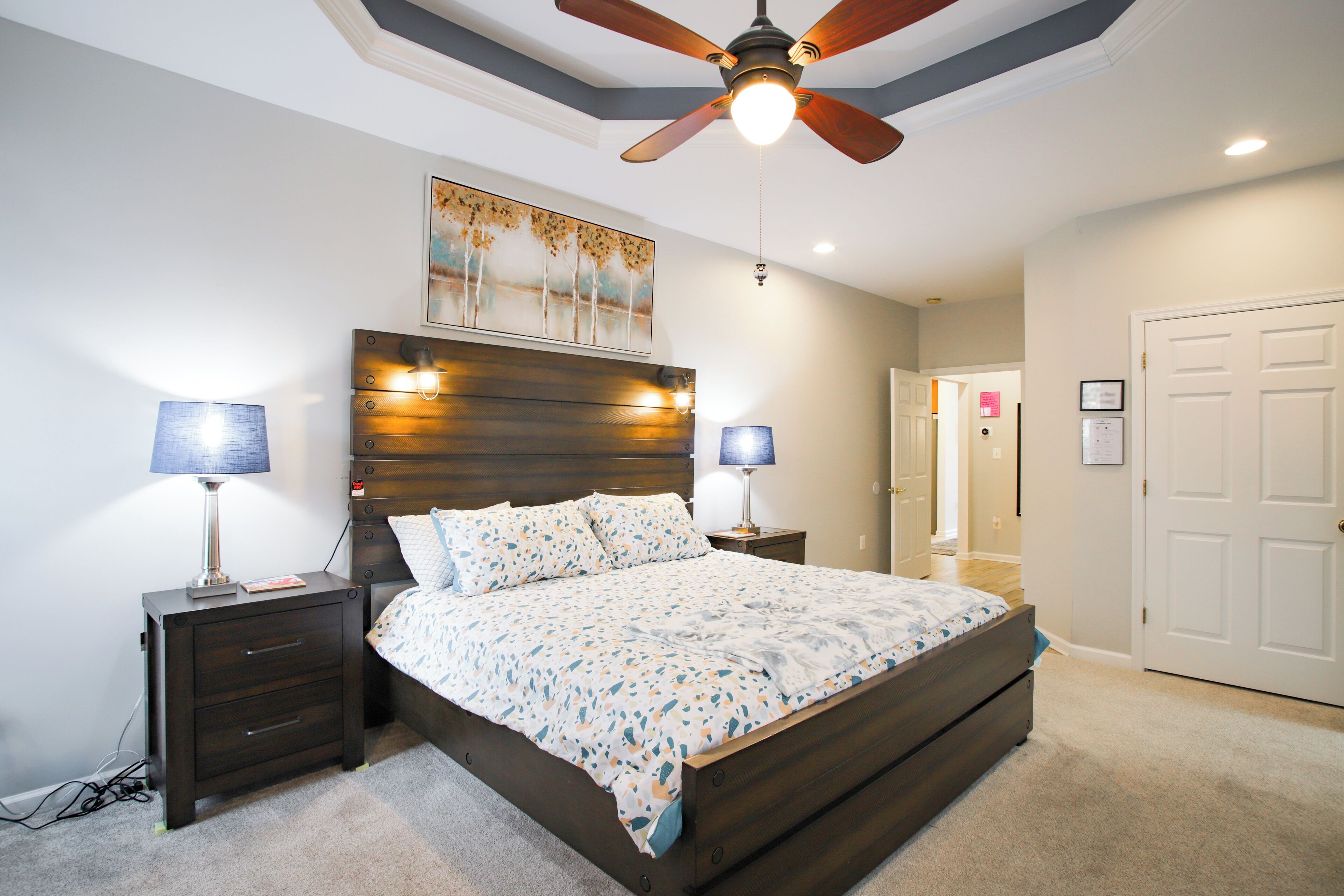 A bedroom with a large bed, two bedside tables with lamps, a ceiling fan, and a door to another room.