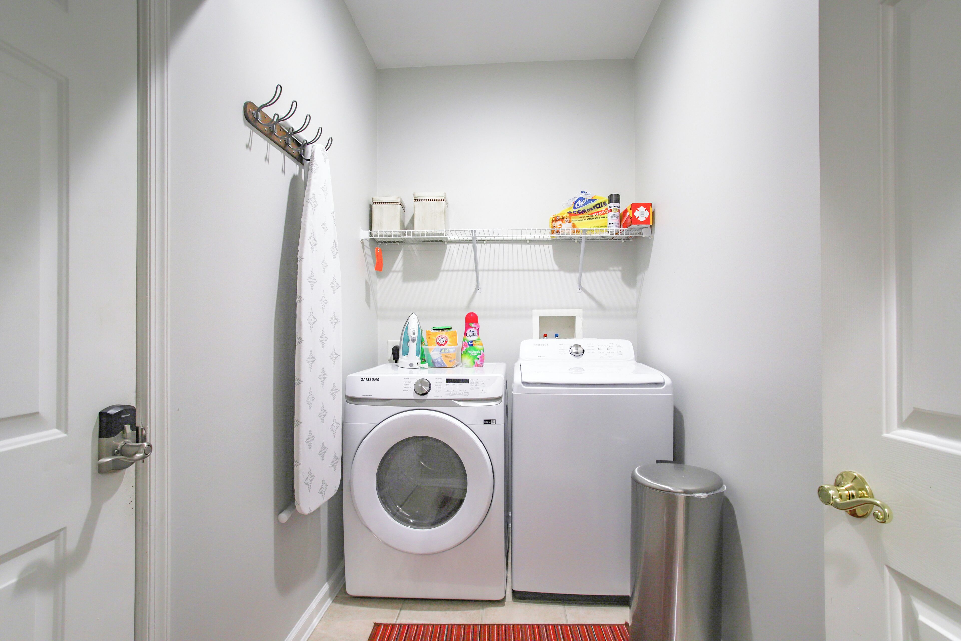Een kleine wasruimte met een wasmachine en droger, een klein plankje met schoonmaakmiddelen en een metalen prullenbak.