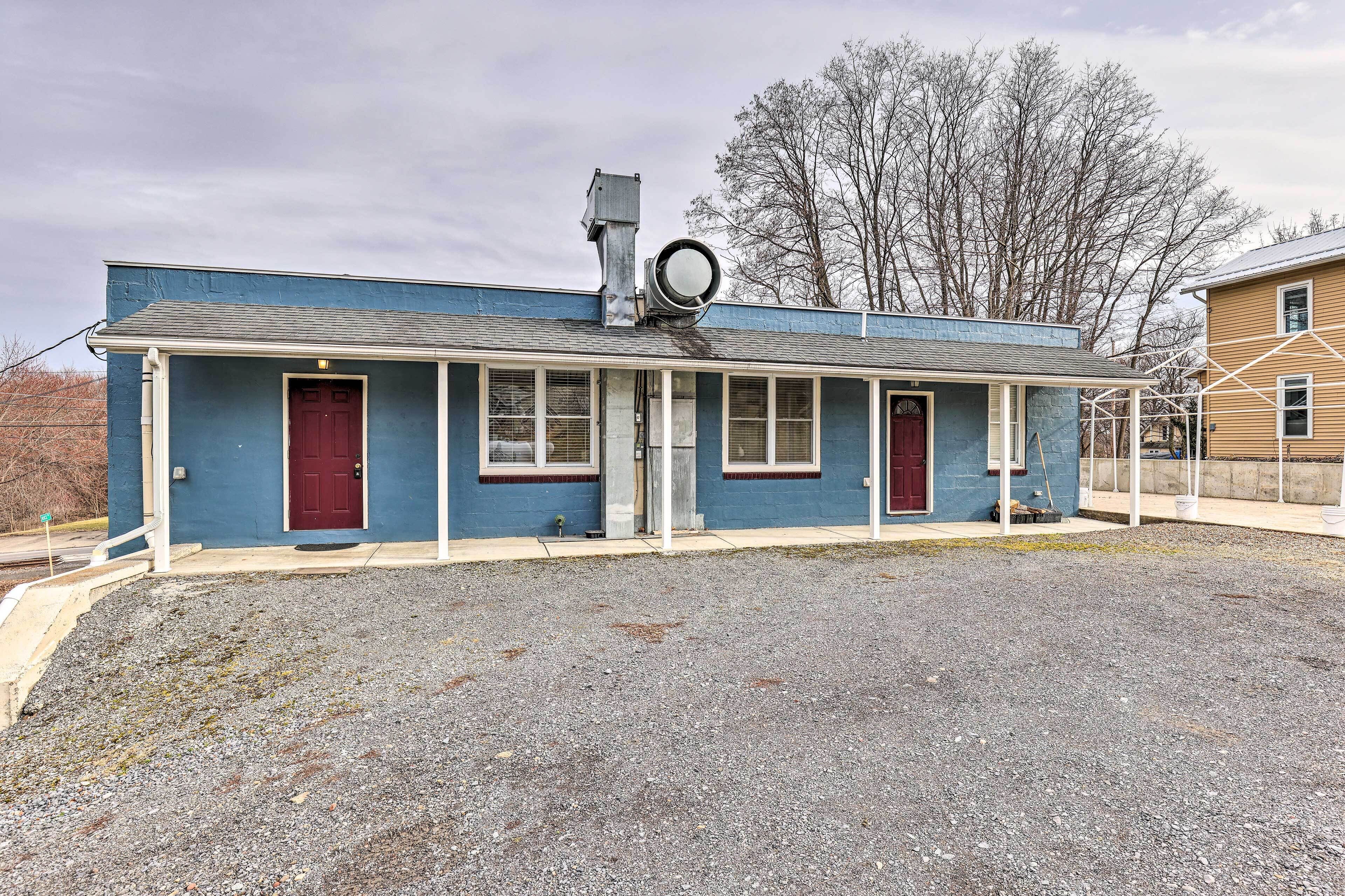 A single-story building with a blue exterior and a red door, featuring a small porch and a gravel driveway.