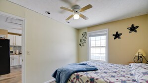 Une chambre à coucher comprenant un lit, un ventilateur de plafond, une fenêtre et une porte donnant sur la cuisine.