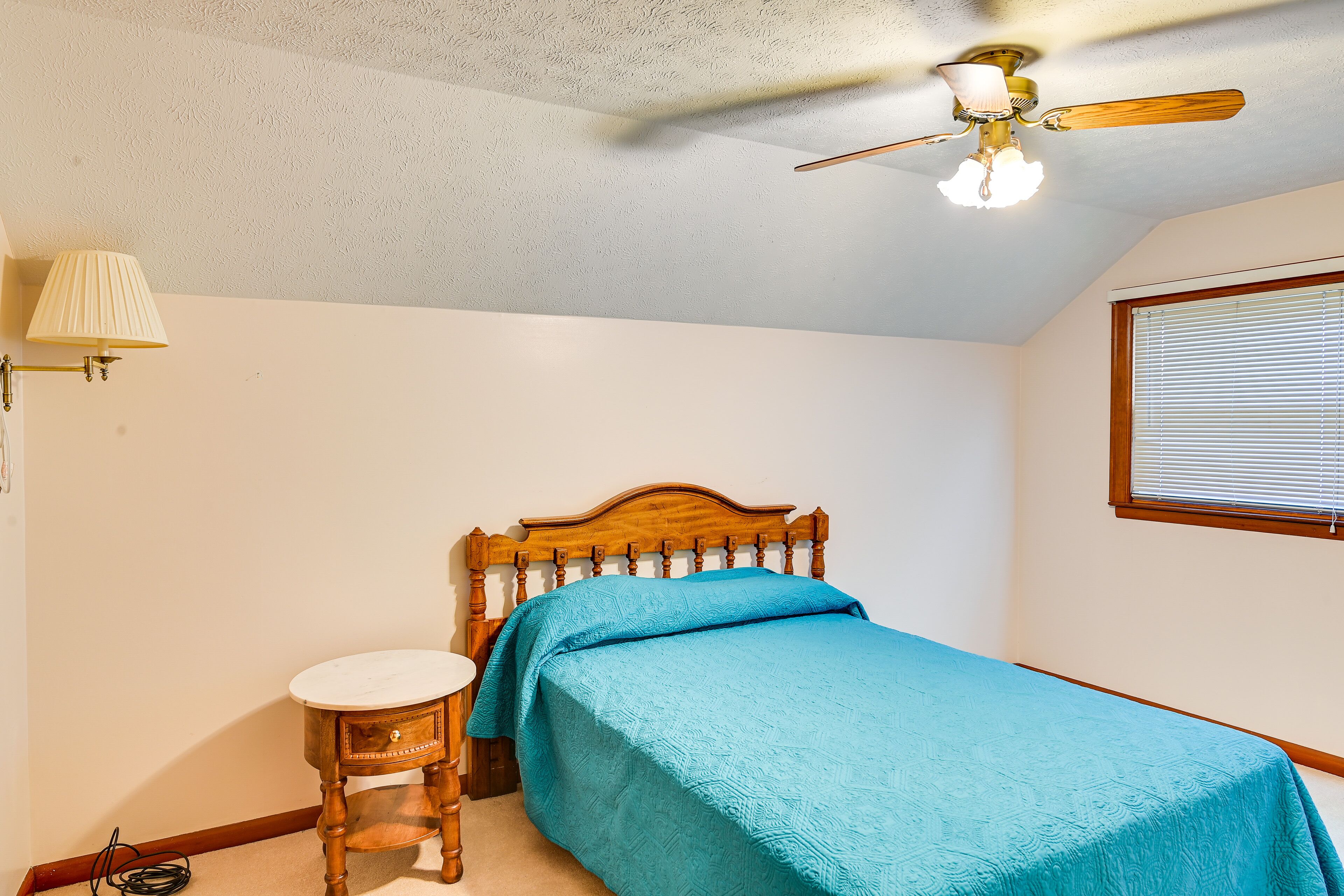 A bedroom with a bed, a wooden nightstand, a ceiling fan, a window with blinds, and a lamp.