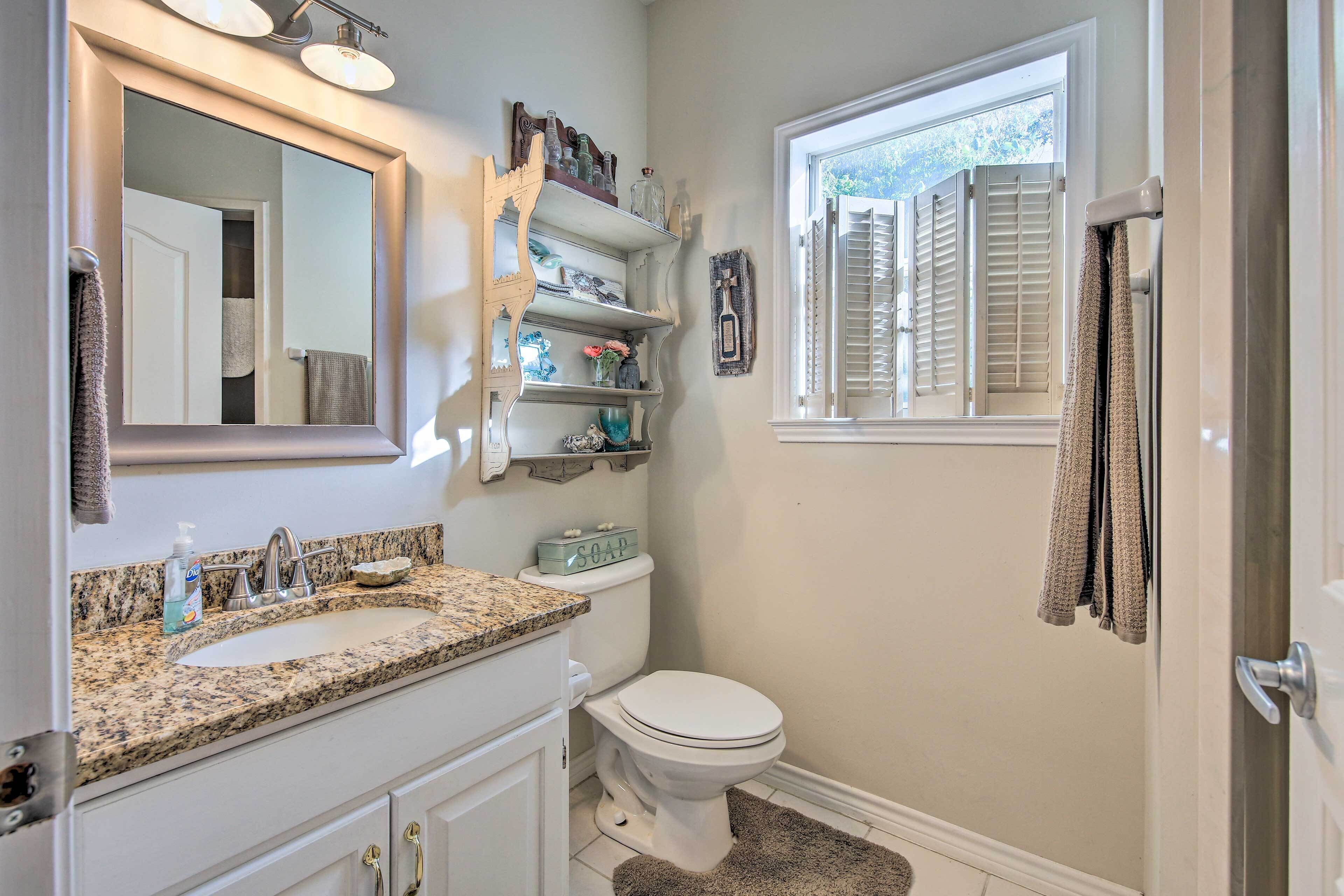 A bathroom with a granite countertop sink, a toilet, a window with shutters, and a mirror with shelves above.