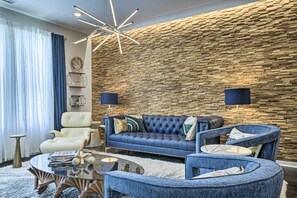 Ein modernes Wohnzimmer mit einem blauen Sofa, einem weißen Sessel und einem runden Couchtisch. Hinter dem Sofa befindet sich eine Wand mit Ziegeloptik und auf beiden Seiten stehen Stehlampen.