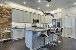 A modern kitchen with white cabinetry, a central island with bar stools, stainless steel appliances, and a unique ceiling light fixture. - Sleek Durham Townhome Near Dtwn w/ Balcony! (Durham)