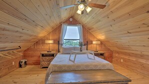 A cozy attic bedroom with a sloped ceiling, a bed with a white bedspread, two bedside tables with lamps, a window with curtains, and a ceiling fan.