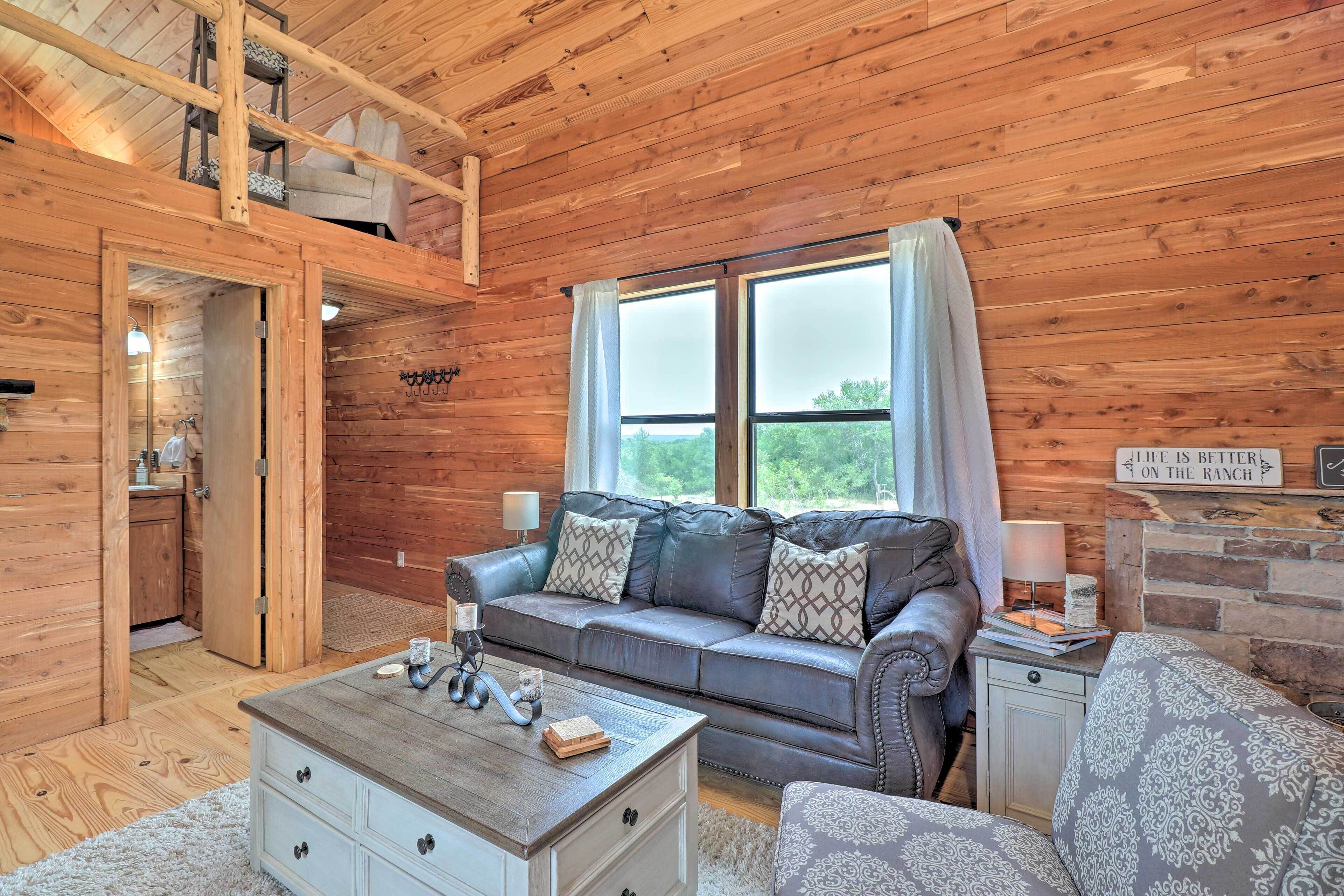 A cozy cabin interior with a wooden ceiling, a sofa with patterned cushions, a coffee table, and a small side table with a lamp.
