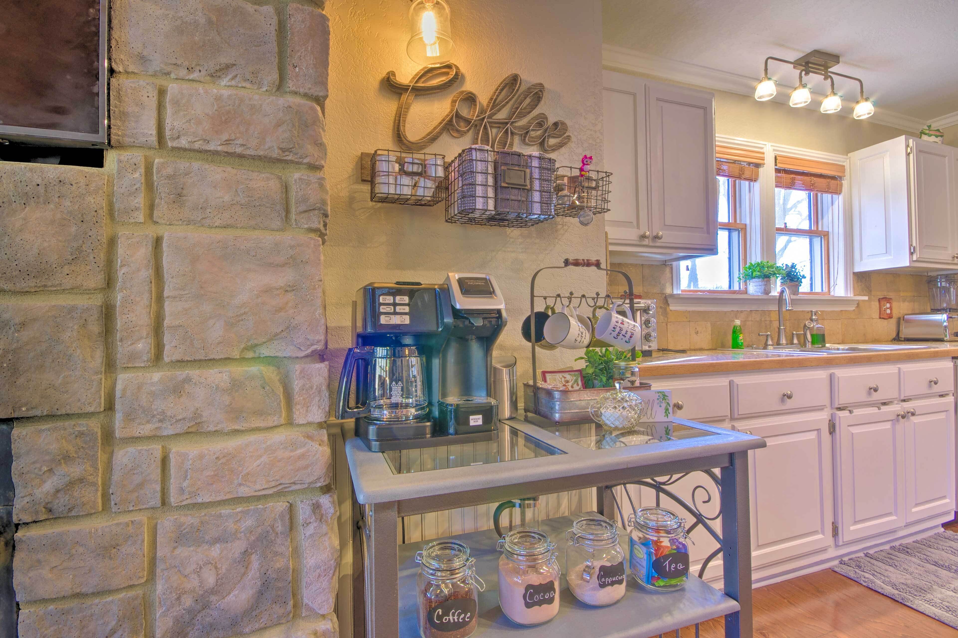 Une cuisine aux murs en pierre, avec un coin café, des meubles blancs et une fenêtre ornée de plantes.