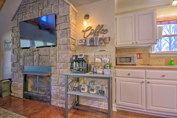 A stone fireplace with a TV mounted above it, a coffee station with jars labeled with coffee types, and a kitchen area with white cabinets and a microwave. - Updated Cabin on White River by Boat Landing! (Calico Rock)