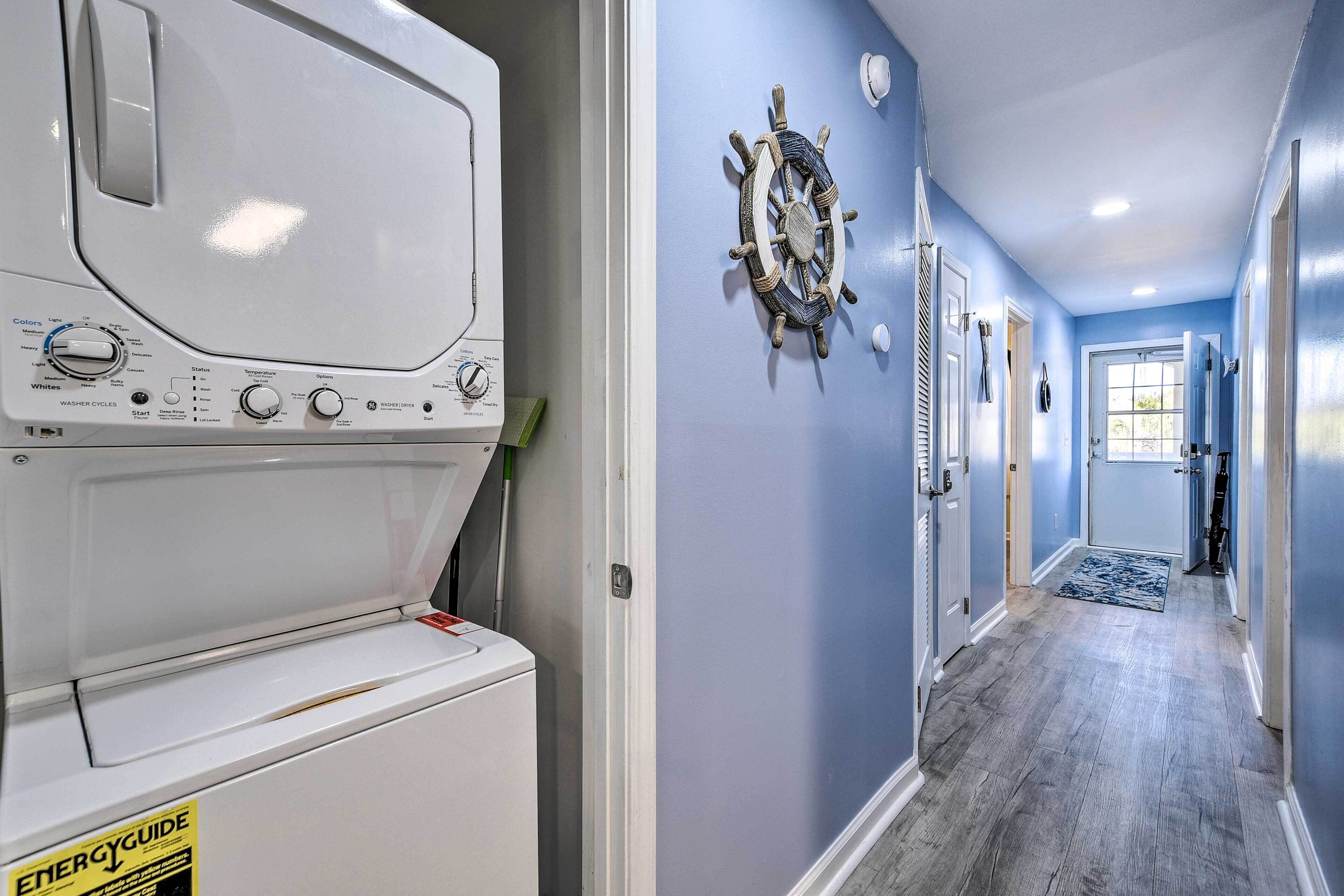 A laundry area with a front-loading washer and dryer, a nautical-themed wall decoration, and a hallway with a wooden floor and blue walls.
