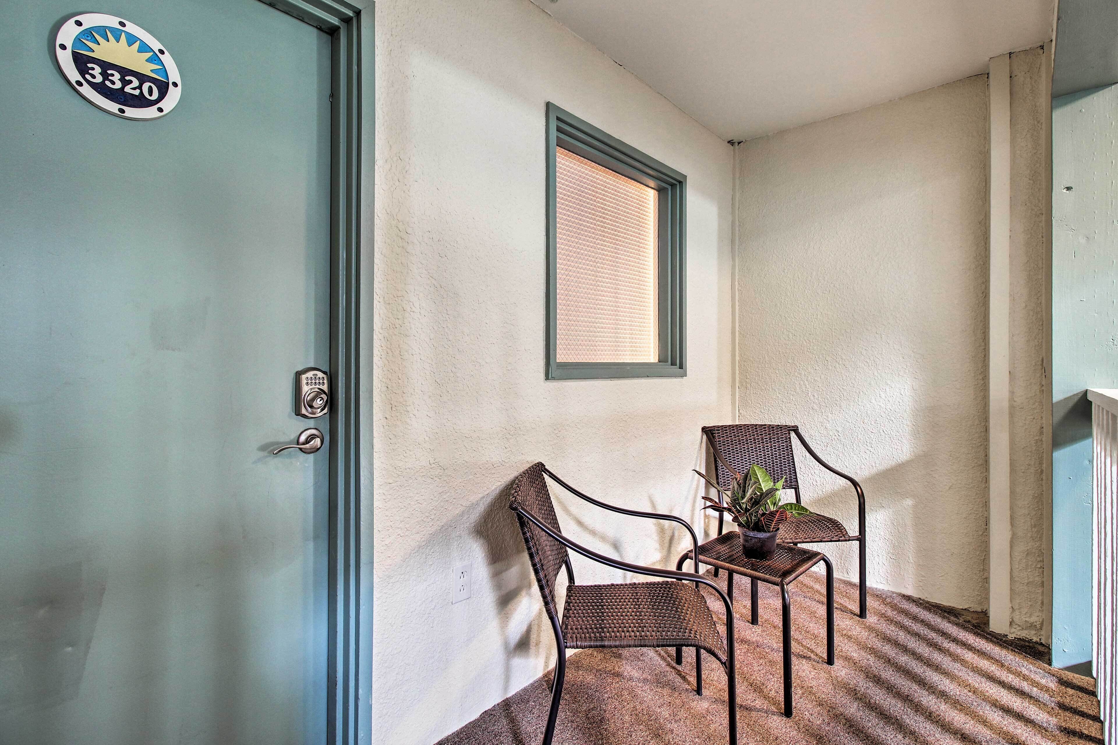 Ein Zimmer mit einer Tür, auf der '3320' steht, einem Fenster mit Jalousien, zwei Korbstühlen und einem kleinen Tisch mit einer Pflanze.