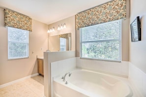 A bathroom with a large bathtub, two windows with blinds, a mirror, and a vanity with a sink.
