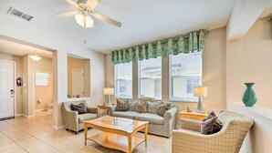 A living room with a beige sofa, a glass coffee table, and patterned armchairs. There are two windows with blinds, a ceiling fan, and a green curtain. - Kissimmee Getaway ~ 4 Mi to Walt Disney World (Kissimmee)