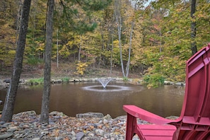 A serene pond with a fountain, surrounded by autumn-colored trees and a red chair in the foreground. - 5 Mi to Raystown Lake Launch: Lucas Mccain Cabin (Huntingdon)