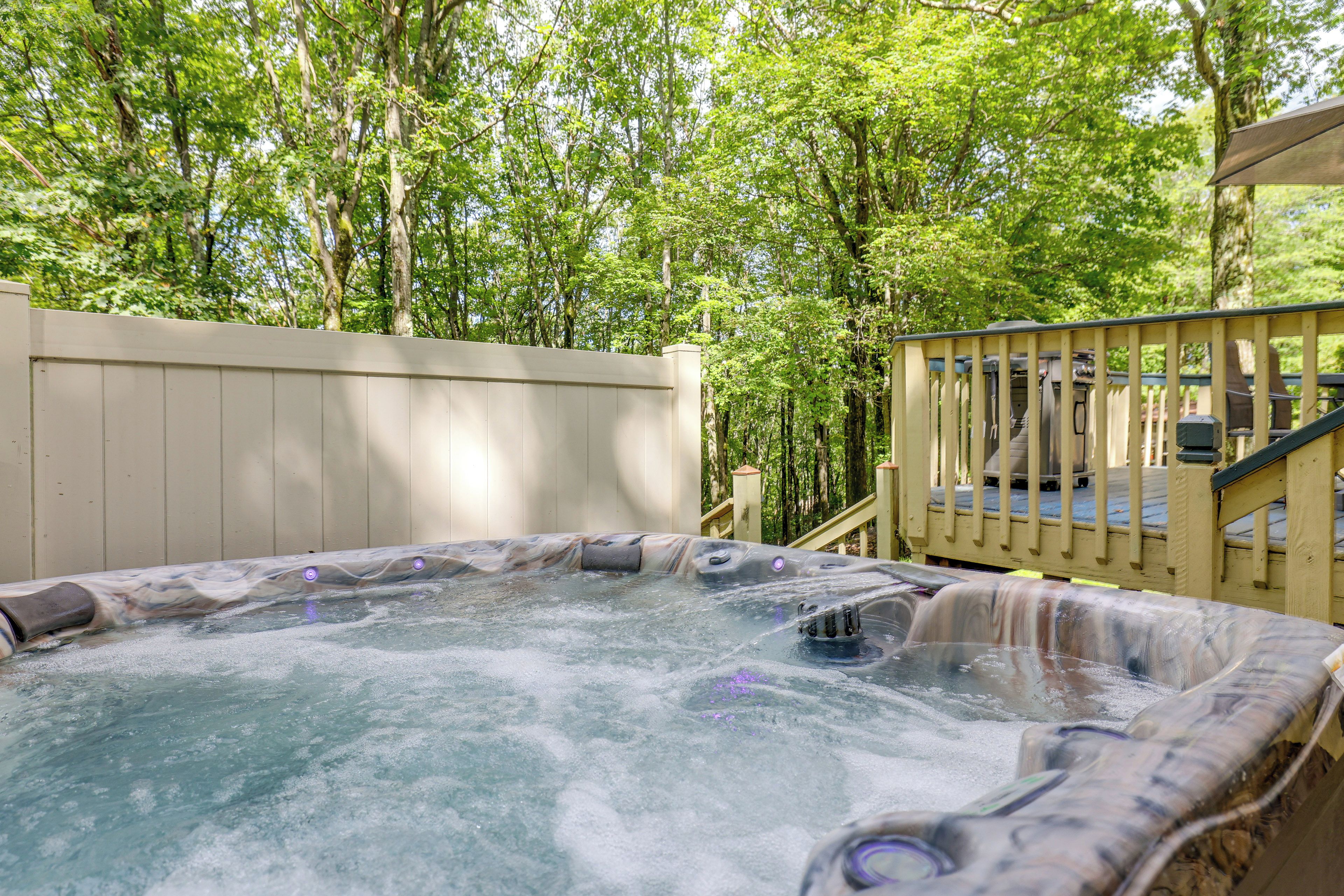 Ein Whirlpool, umgeben von einer Holzterrasse und einem Holenzaun, mit Bäumen im Hintergrund.