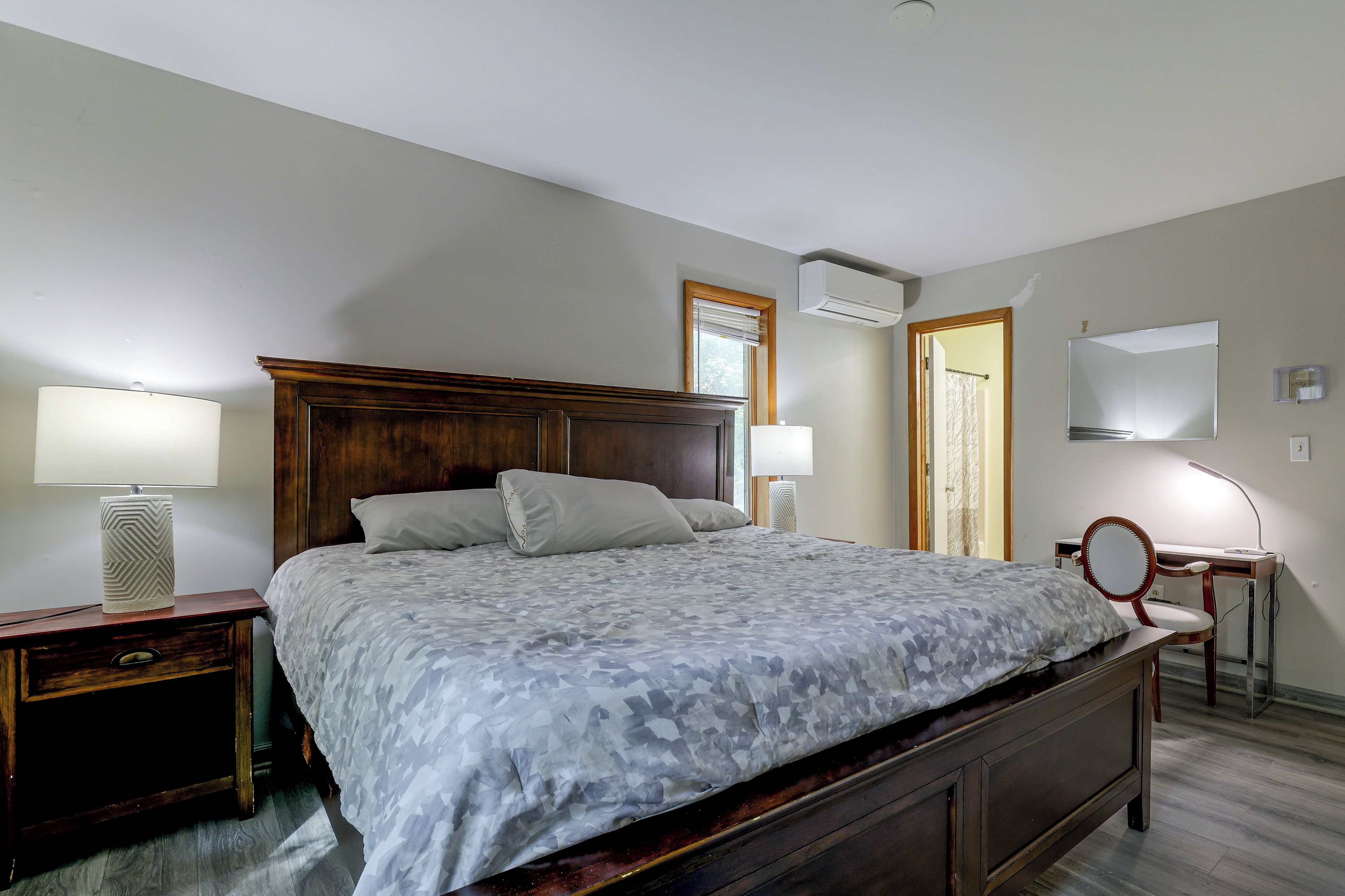 A bedroom with a large bed, two bedside tables, a desk with a chair, a mirror, and an air conditioning unit.