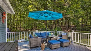A deck with wicker furniture, a blue umbrella, and a view of a forest. - Stunning South Fallsburg Home w/ Pool & Deck! (South Fallsburg)