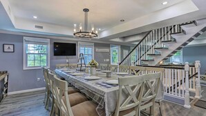 A dining area with a long table, chairs, and a chandelier. There is a staircase with a railing leading to an upper level. - Stunning South Fallsburg Home w/ Pool & Deck! (South Fallsburg)