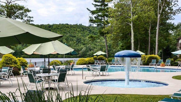 An outdoor pool area with lounge chairs and umbrellas, surrounded by greenery and a view of a lake. - 1 Mi to Big Boulder: Lakefront Unit w/ Pool Access (Lake Harmony)