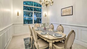 Un comedor con una mesa grande dispuesta para seis personas, rodeada por seis sillas. Encima de la mesa hay una araña, y una ventana con vistas a árboles.