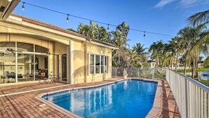 Una piscina con solárium empedrado, un porche cubierto con mobiliario exterior y vistas a palmeras y una casa al fondo.
