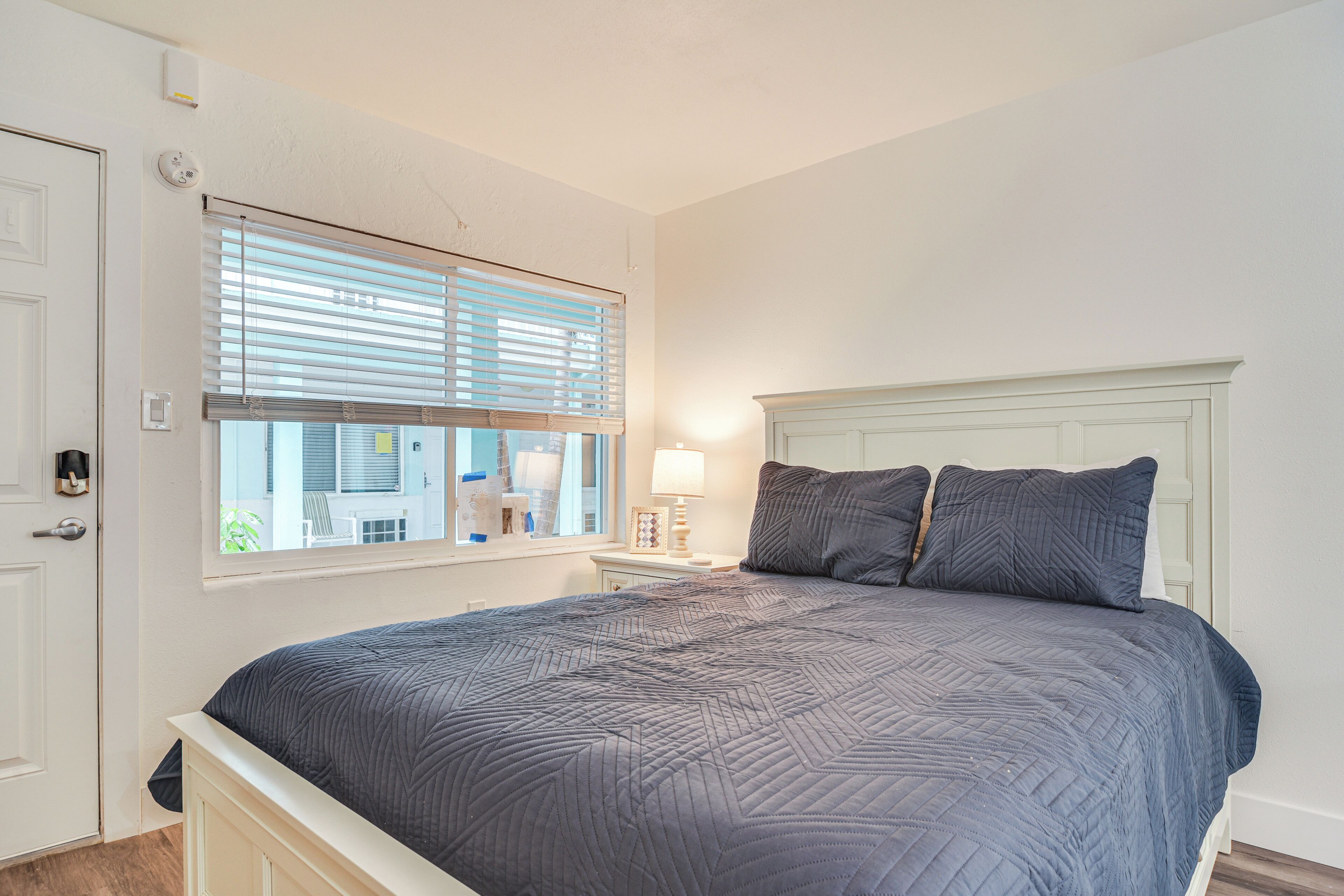 A bedroom with a bed, a nightstand, a lamp, a window with blinds, and a door.