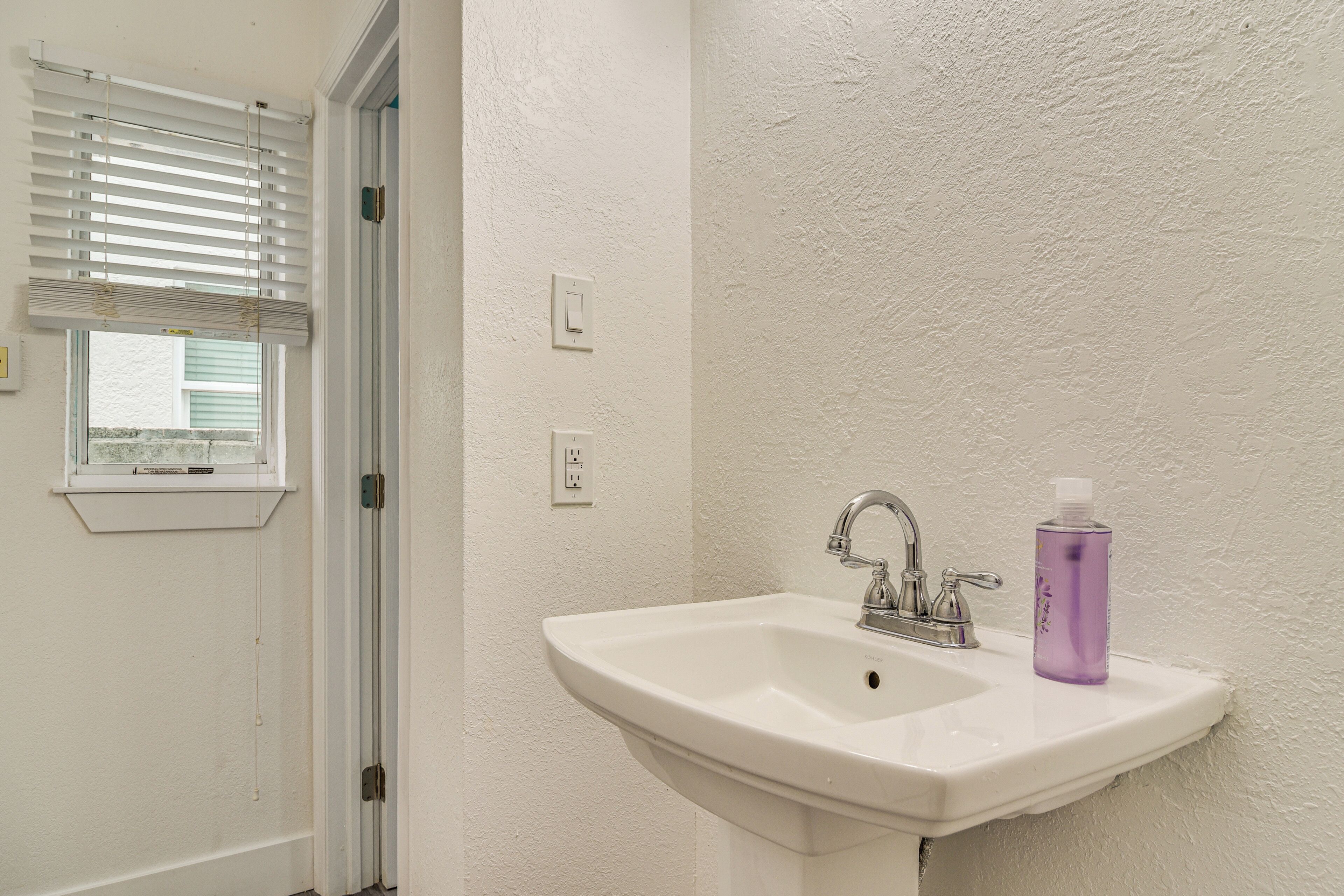 A bathroom with a white sink, a window with blinds, and a purple bottle on the sink.