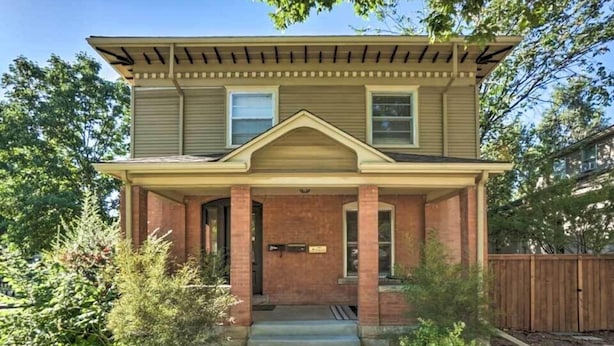 Una casa a due piani con un portico anteriore, facciata in mattoni e un piccolo giardino con arbusti.