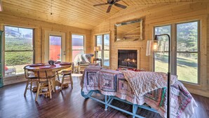 A cozy cabin interior with a wooden ceiling, a fireplace, a bed with a quilt, a dining table with chairs, and large windows offering a view of the outdoors. - Nature Escape in Wytheville w/ Covered Porch! (Speedwell)