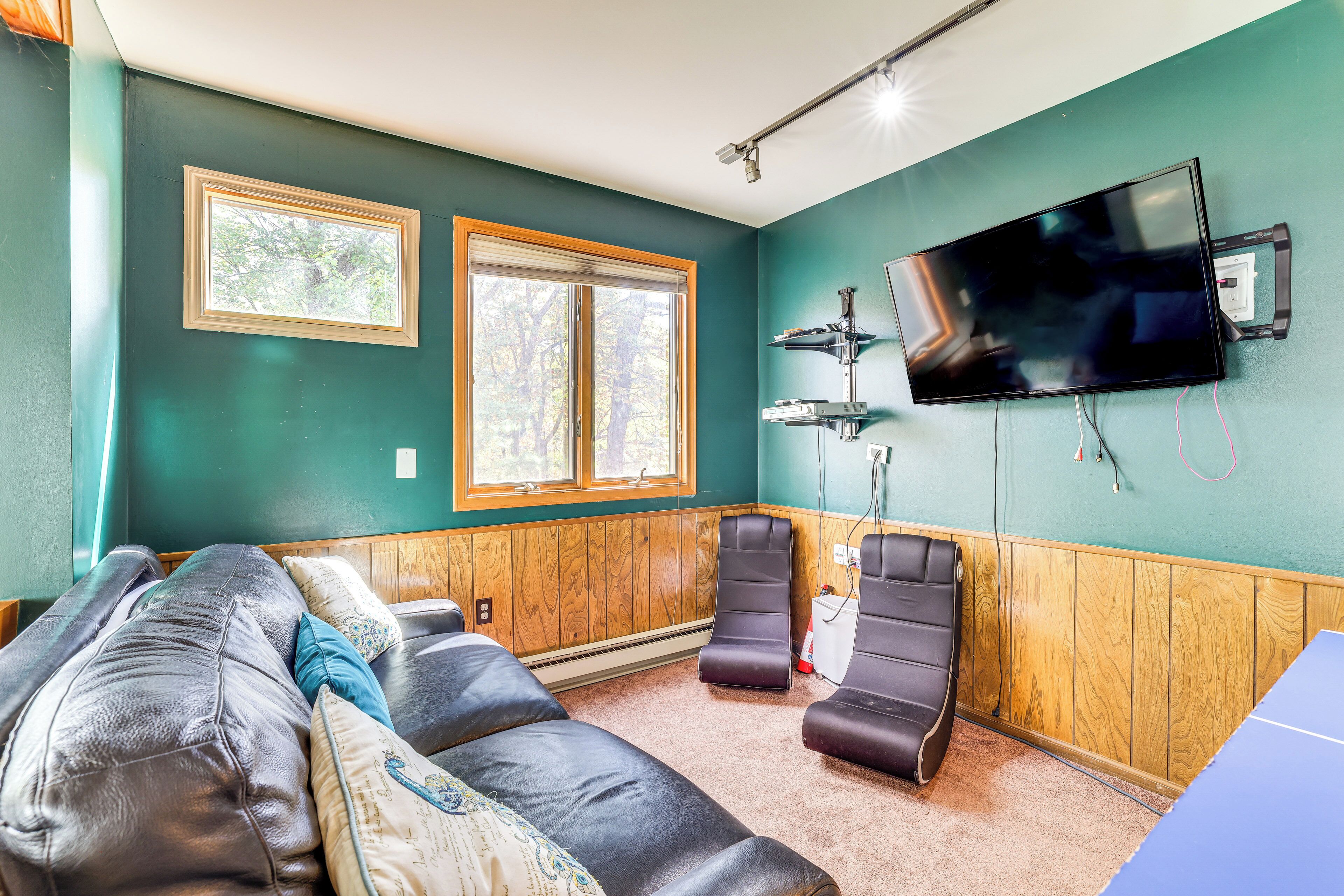 A room with a large flat-screen TV mounted on the wall, a wooden paneled wall, two windows, a massage chair, and a massage table.